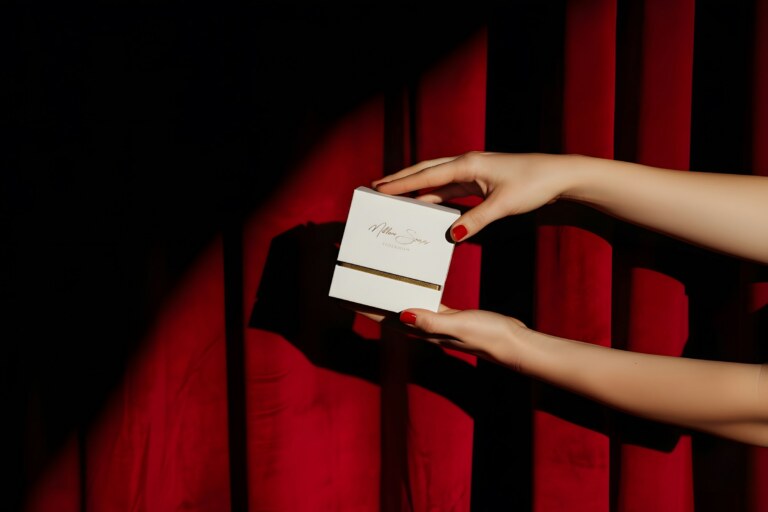Hands holding a small white gift box against a red curtain